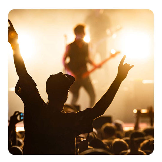 Image of a person holding up his arms in a concert event space, with a person playing guitar on the stage in front of him.