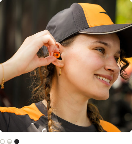 A close-up lifestyle shot of a woman wearing a black and orange baseball cap and a matching black and orange shirt, wearing McLaren x Loop Switch 2 earplugs